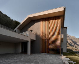 Exterior facade of a contemporary private residence in Ticino, featuring a full-height vertical timber louvre screen in warm cedar tones. The wooden slats catch direct afternoon sunlight against a rendered concrete structure, with alpine rock face visible in the background. 