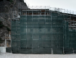Full-height scaffolding draped in dark green mesh netting at a construction site in Limone sul Garda, with a steep limestone cliff rising directly behind. Timber roof structure and upper floor framing visible above the netting. Gravel foreground, overcast sky. 