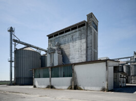 Industrial grain storage facility of CO M.A. Cer in the Po Valley near Parma, featuring a weathered concrete elevator tower with faded lettering alongside corrugated steel silos and conveyor structures. Flat agricultural landscape visible on the horizon under an open sky. 