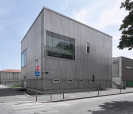 xterior corner view of Gymnase René Lacoste in Troyes, a compact cubic sports hall clad entirely in corrugated metal sheeting. Large recessed glazing on the upper level reflects the surrounding urban context. The building appears to float above street level, with basement ventilation slots visible at the base. 