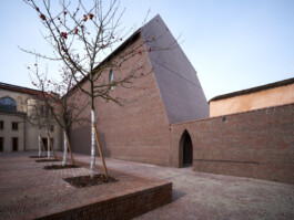 Courtyard of the Musée Unterlinden extension in Colmar, designed by Herzog & de Meuron. New brick volume with a sharply angled copper roof rises between historic rendered walls. Bare-branched trees with red fruit line the brick-paved courtyard in late autumn light. 