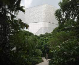 Die geschwungene, schuppenartige Aluminiumfassade von Central Embassy in Bangkok erhebt sich über dichtem tropischem Grün; auf einem Gartenweg gibt eine einzelne Figur den Massstab. 