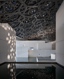 Innenraum des Louvre Abu Dhabi unter der perforierten Kuppel: gefiltertes Sonnenlicht («Rain of Light») fällt auf weisse Kubaturen und ein Wasserbecken, eine einzelne Figur in Weiss gibt den Massstab. 