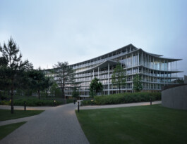 Geschwungener, weisser Hauptsitz von Lombard Odier in der blauen Stunde, mit schlanken Stützen und auskragenden Geschossen, umgeben von Parklandschaft – Bellevue, Genf. 